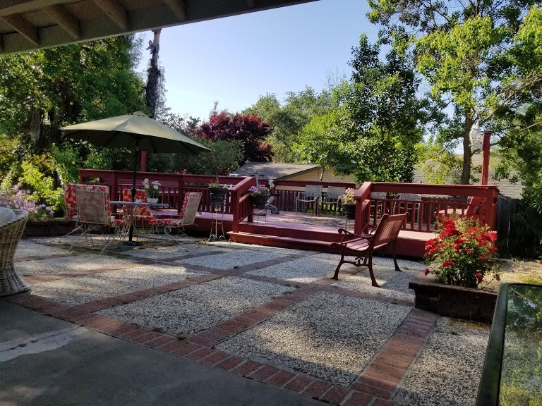 A sunny backyard patio with a wooden deck and outdoor furniture surrounded by trees and plants.