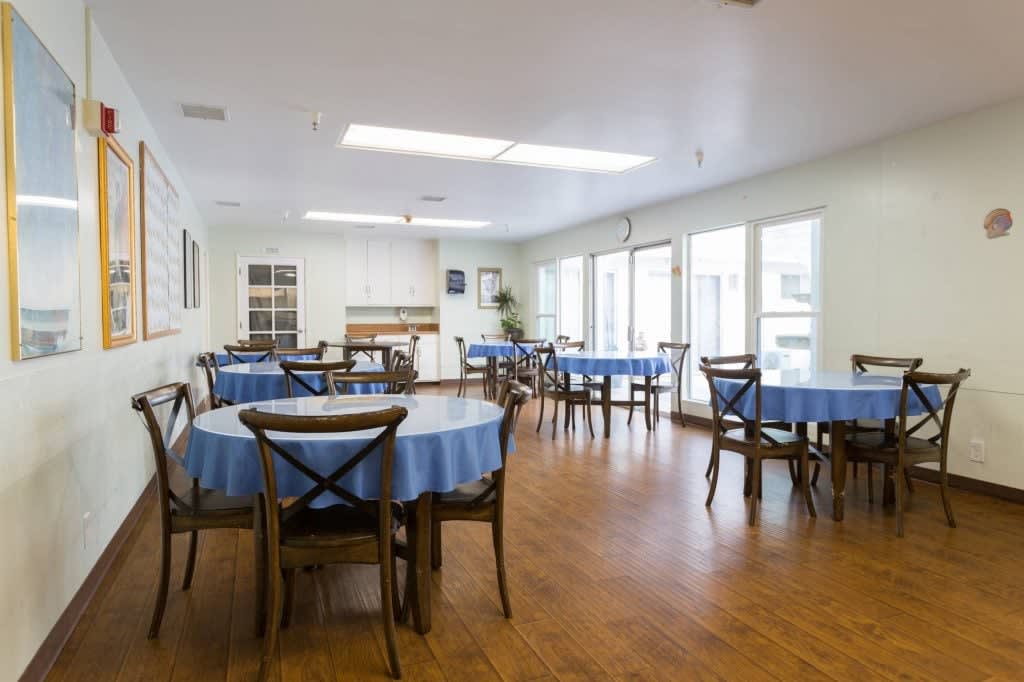 A brightly lit dining room with multiple tables covered in blue tablecloths and wooden chairs.