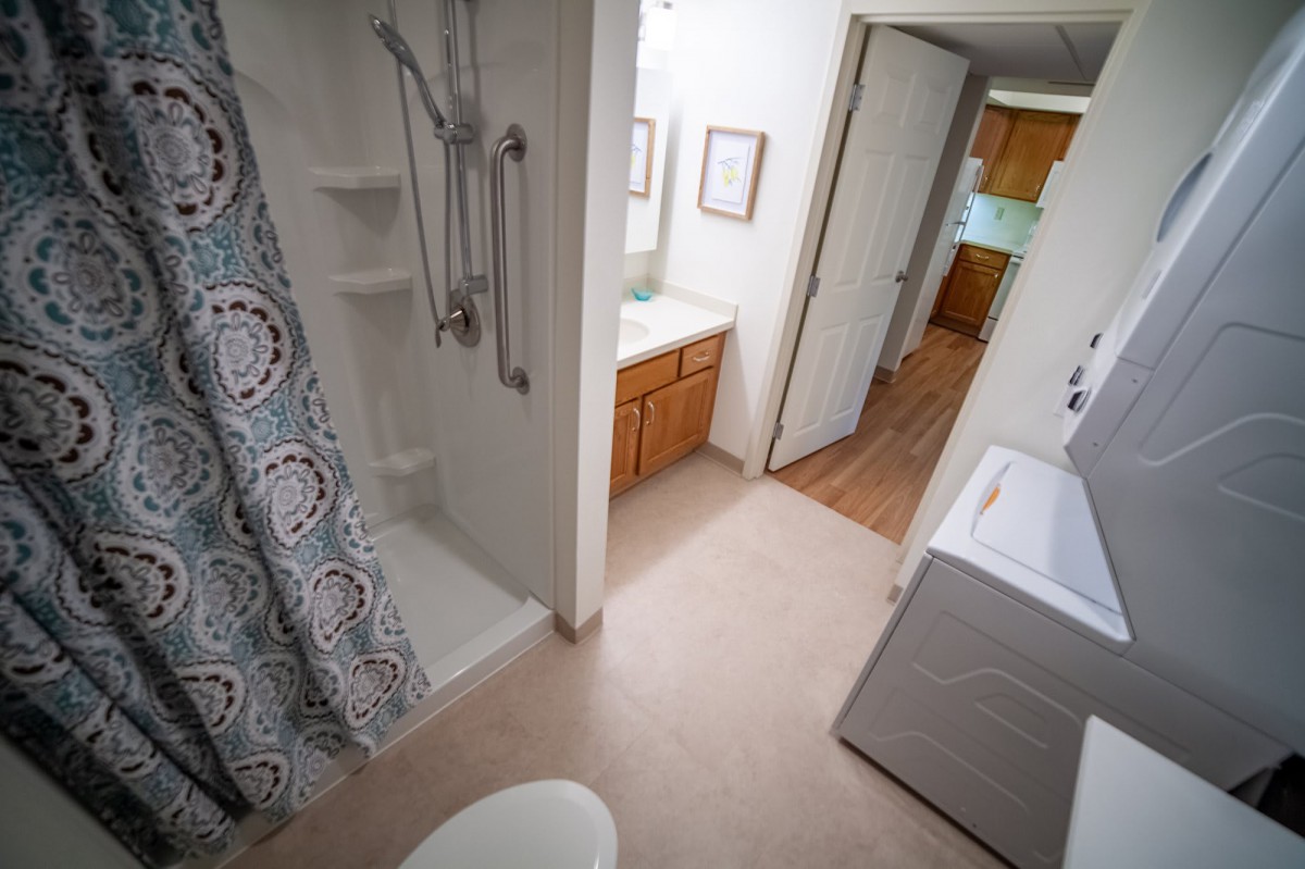 A home bathroom with a shower, washer, and hallway leading to the kitchen.