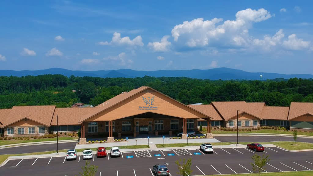 A school building with a parking lot, surrounded by trees and mountains under a blue sky with clouds.