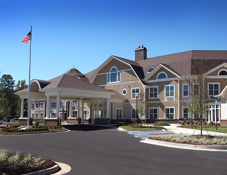 A large residential building with a driveway and an American flag in front, surrounded by landscaping under a clear blue sky.