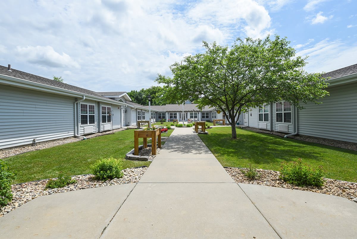 A peaceful courtyard surrounded by buildings with a pathway, benches, and lush greenery under a cloudy sky.