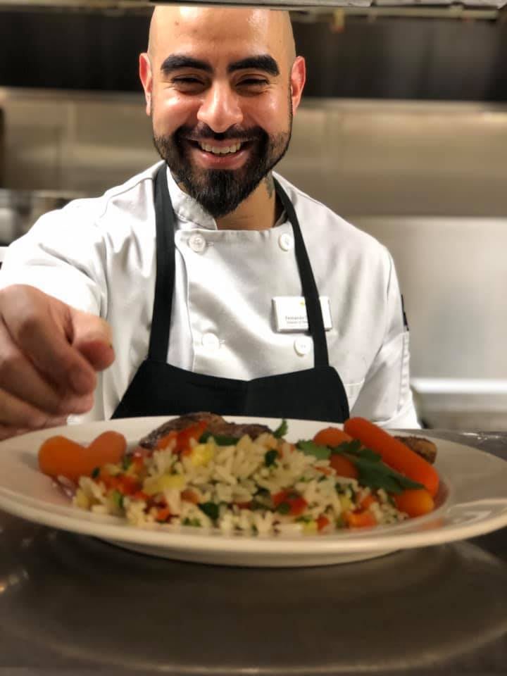 A smiling chef presenting a beautifully garnished plate of food in a kitchen.