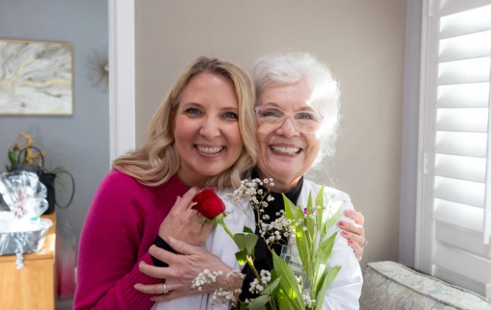 A younger and an elderly woman smiling together, holding flowers indoors.