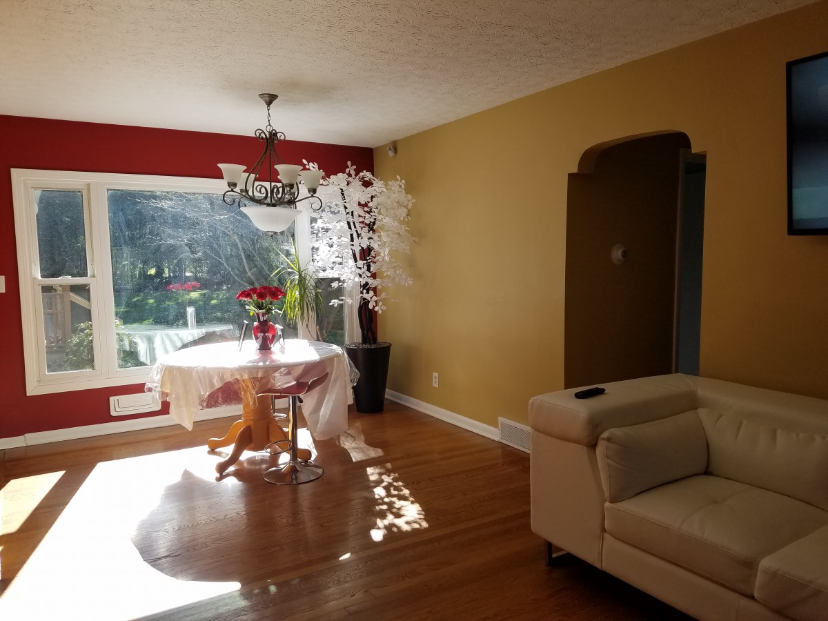 A cozy living and dining area featuring a round table with chairs, a chandelier, a couch, and large windows allowing natural sunlight to brighten the space.