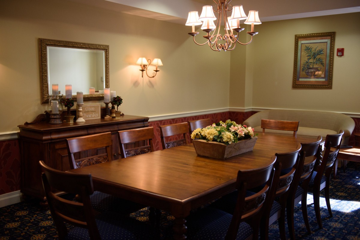 A dining room with a wooden table and chairs, featuring a floral centerpiece and chandelier lighting.