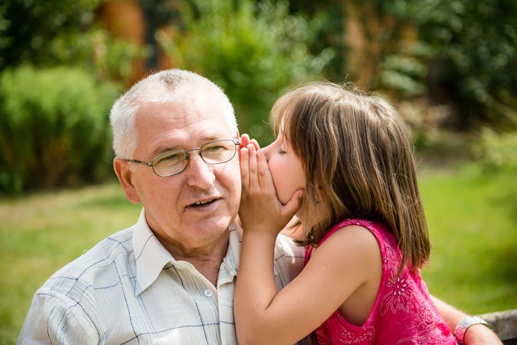A young girl whispers a secret to her grandfather while sitting outdoors.