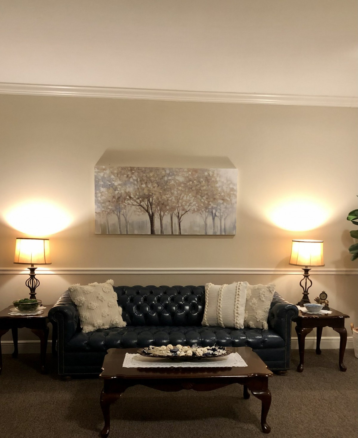 A cozy living room with a blue sofa, illuminated by two lamps, adorned with pillows and artwork of trees on the wall.