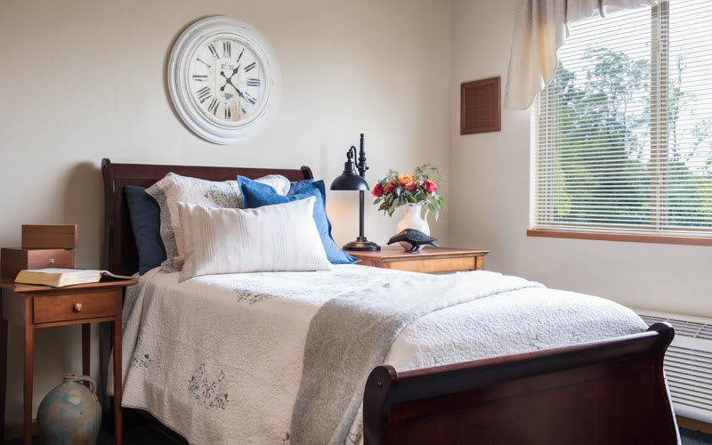 A cozy bedroom with a neatly made bed, adorned with decorative pillows and a nearby nightstand featuring a lamp and flowers.
