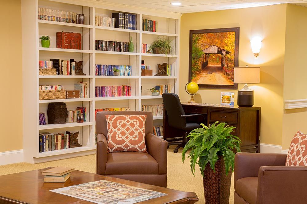 A cozy reading room with bookshelves lined with books, a comfortable armchair, and a desk beneath a colorful painting.