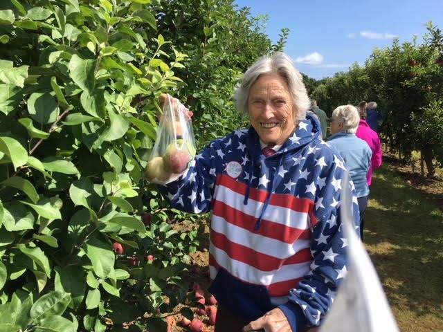 A woman wearing a flag-patterned jacket is apple picking in a garden.
