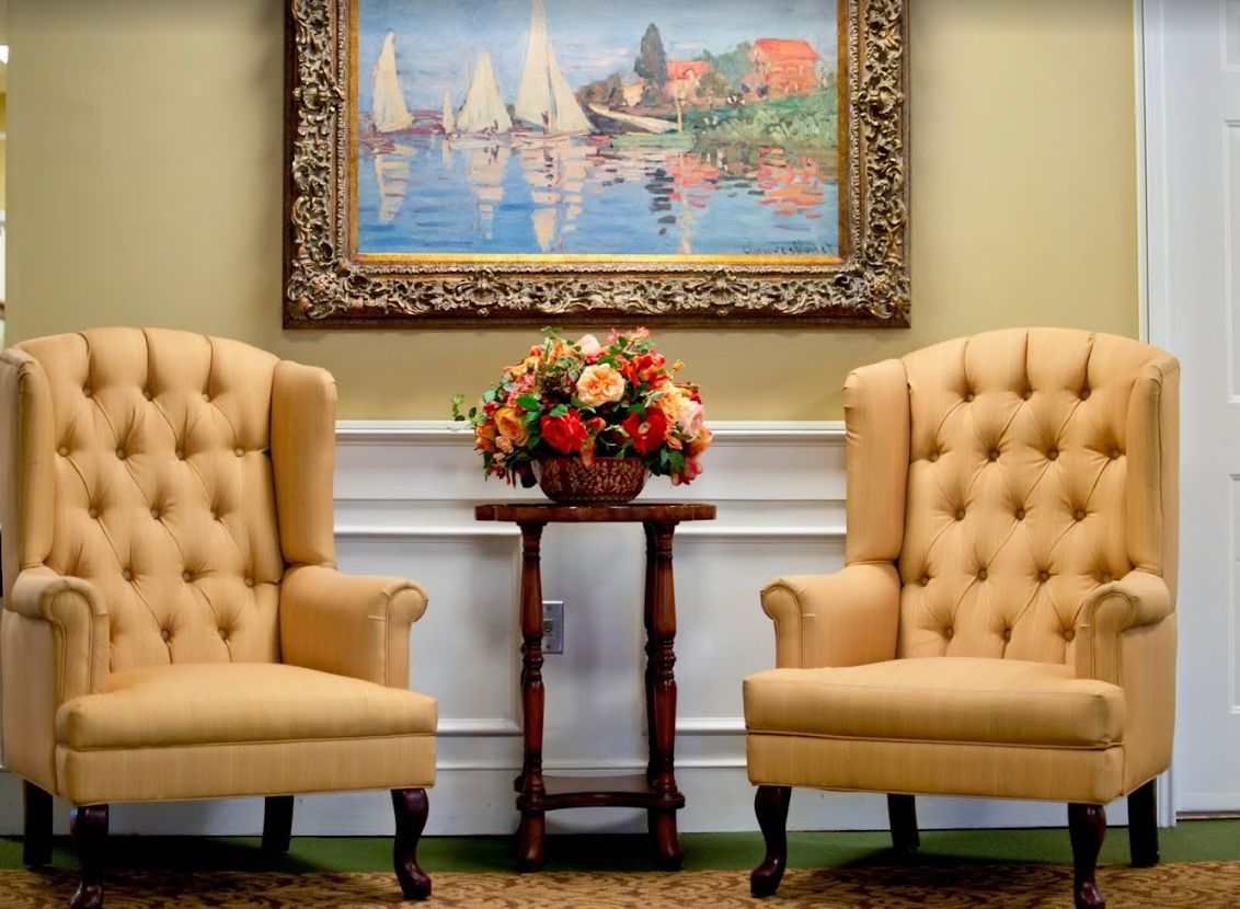 A cozy setting with two upholstered chairs and a floral arrangement, set against a backdrop of a framed painting depicting sailboats on water.
