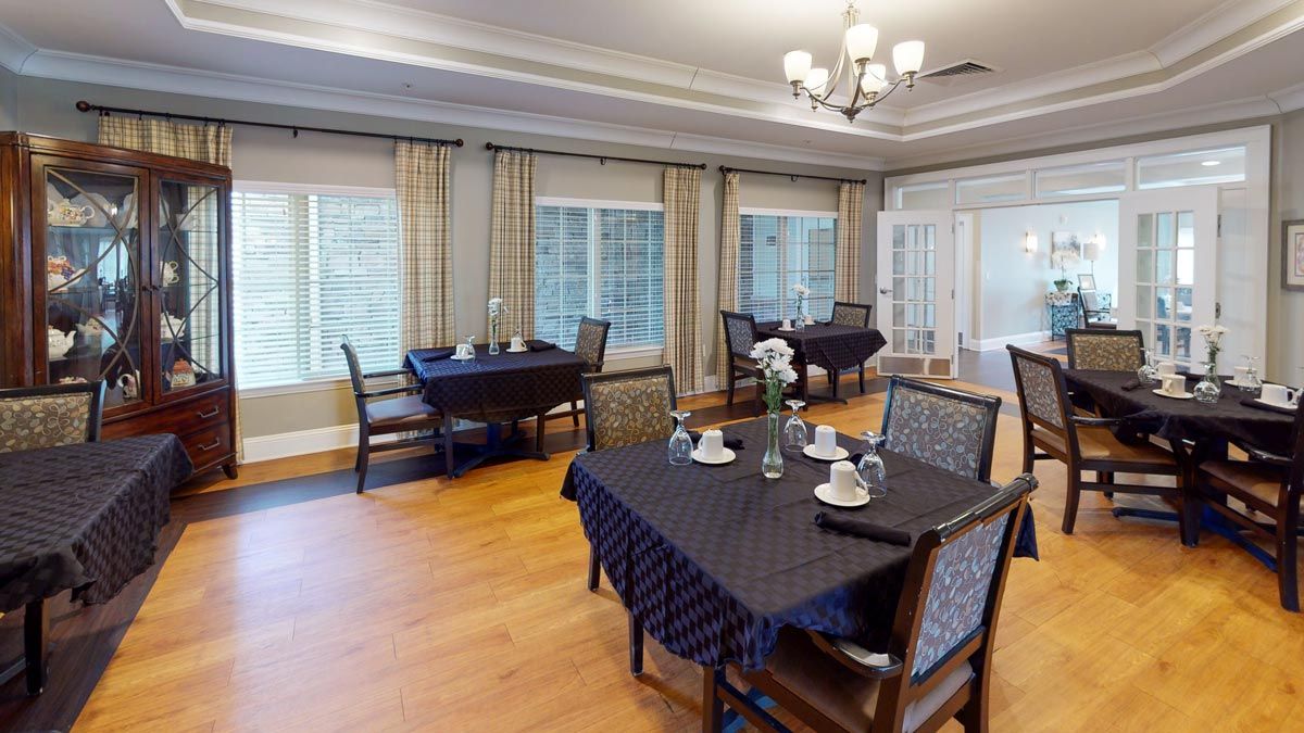 A well-appointed dining room with neatly arranged tables and chairs, featuring large windows, elegant curtains, and a chandelier above a hardwood floor.