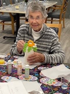 An elderly woman is engaged in a crafting activity, working on an art project at a table filled with art supplies.