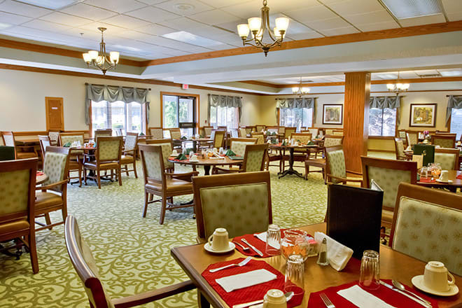A cozy dining room with elegantly set tables and comfortable chairs, illuminated by chandeliers.