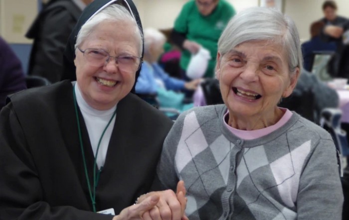 Two elderly women, one a nun, smiling and holding hands in a warm show of friendship.