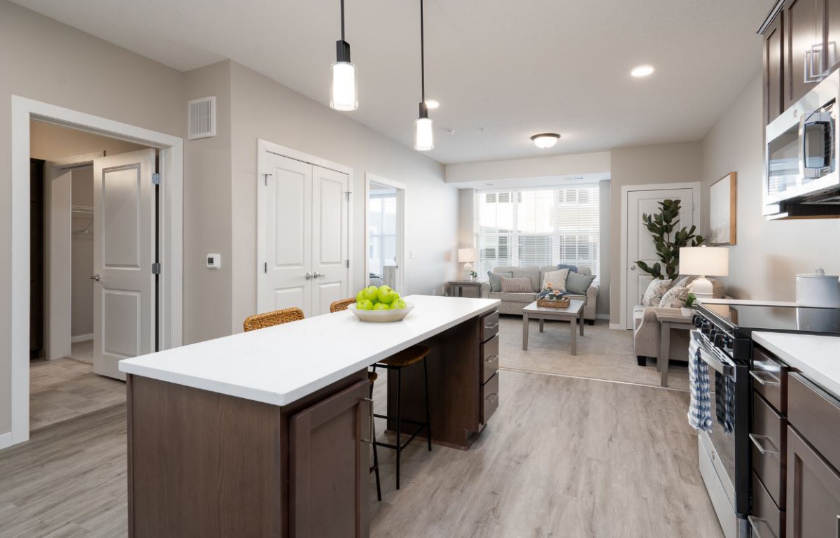 A modern kitchen with an island counter opens into a cozy living area, featuring minimalist interior design and ample natural light.
