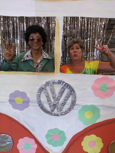 Two individuals dressed in vibrant 70s-style costumes pose playfully behind a decorated Volkswagen-themed backdrop.