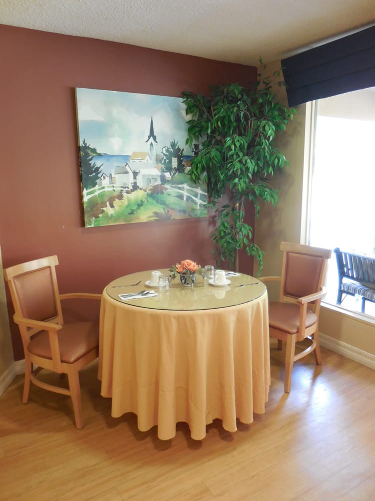 A cozy dining nook with a round table set for two, adorned with a peach tablecloth and complemented by elegant chairs. An indoor plant and a landscape painting enhance the atmosphere, while natural light streams in through a nearby window.