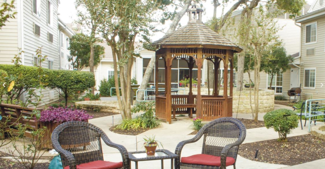 A tranquil garden setting featuring a wooden gazebo with surrounding chairs and lush greenery.