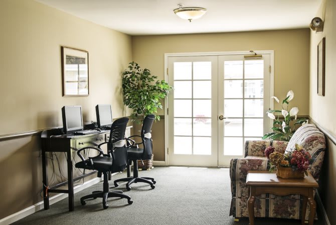 A small office space with computers, chairs, and plants near large windows.