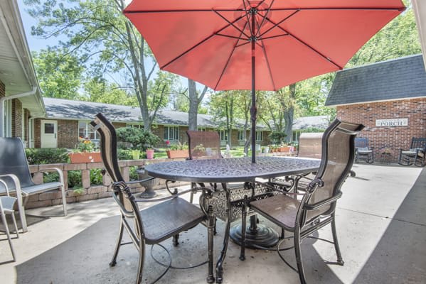 A cozy patio setup with a table, chairs, and a red umbrella in a peaceful garden setting.