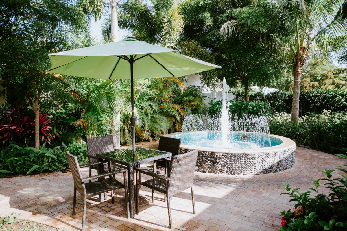 A tranquil patio seating area with a green umbrella and a nearby fountain in a lush garden setting.