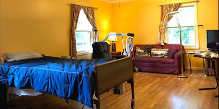 A cozy room with a hospital bed and a sofa, decorated with yellow walls and windows with curtains.