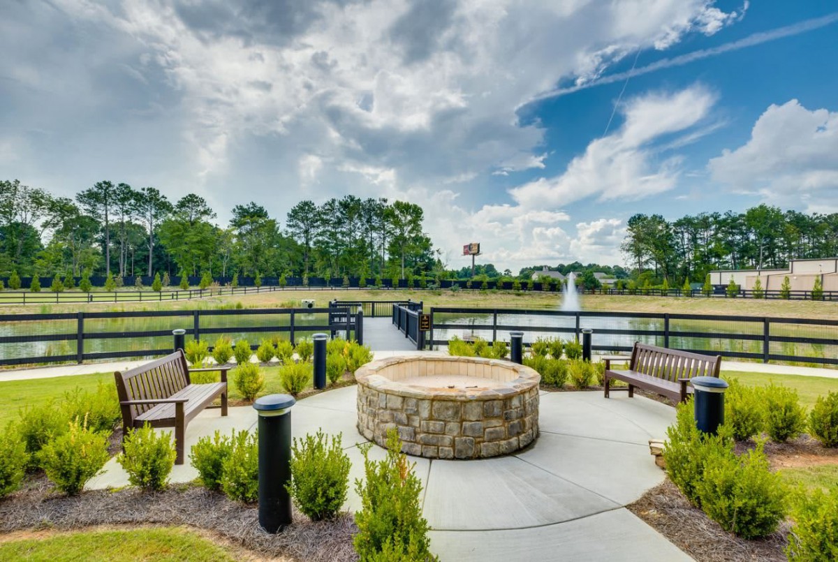 A scenic outdoor area featuring a stone fire pit surrounded by benches, overlooking a pond with a fountain under a cloudy sky.