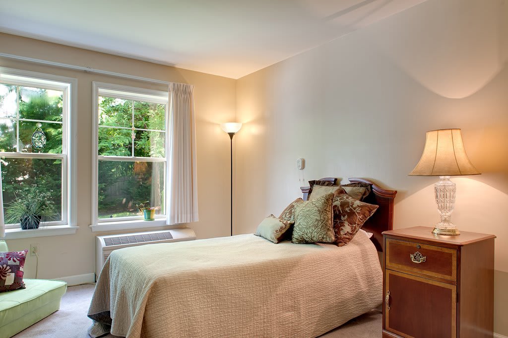 A cozy bedroom featuring a single bed, a window with curtains, and ambient lighting from a floor lamp.