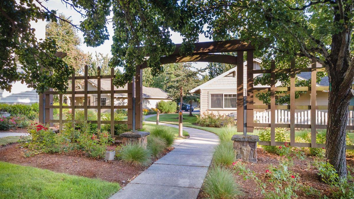 A charming garden pathway leads under an archway towards a house, surrounded by lush greenery and landscaping.