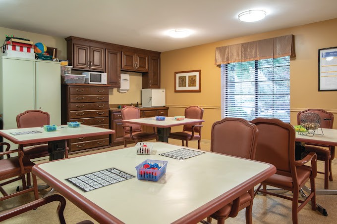 A cozy room with multiple tables and chairs, featuring a kitchenette area with cabinets and a window.