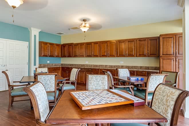 A cozy game room with tables and chairs, featuring a kitchenette and ceiling fan.