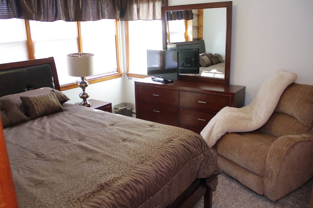 A cozy bedroom featuring a bed, dresser with a mirror, TV, and other furnishings.