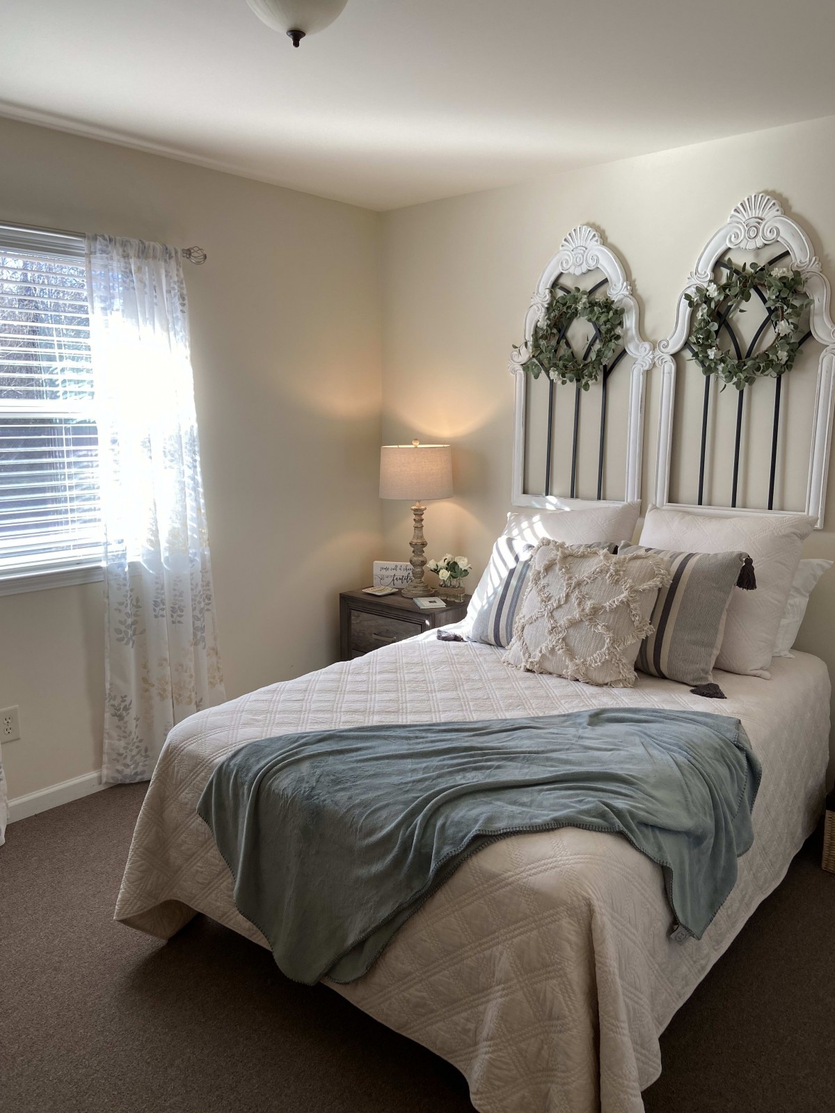 A cozy and well-decorated bedroom with a bed adorned with pillows and a decorative headboard, gently lit by natural light from the window and a lamp on the nightstand.