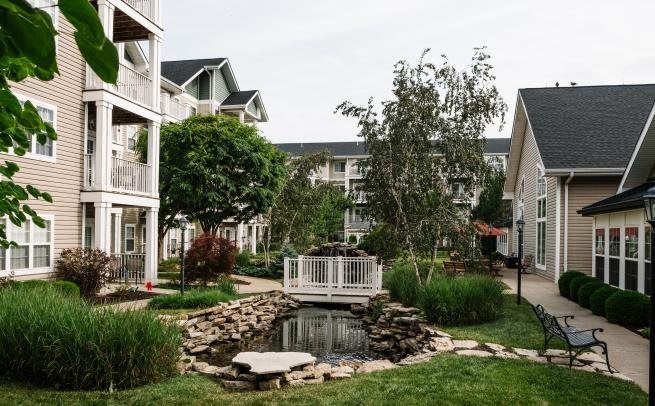 A serene courtyard in an apartment complex featuring a pond and a small bridge surrounded by lush greenery.