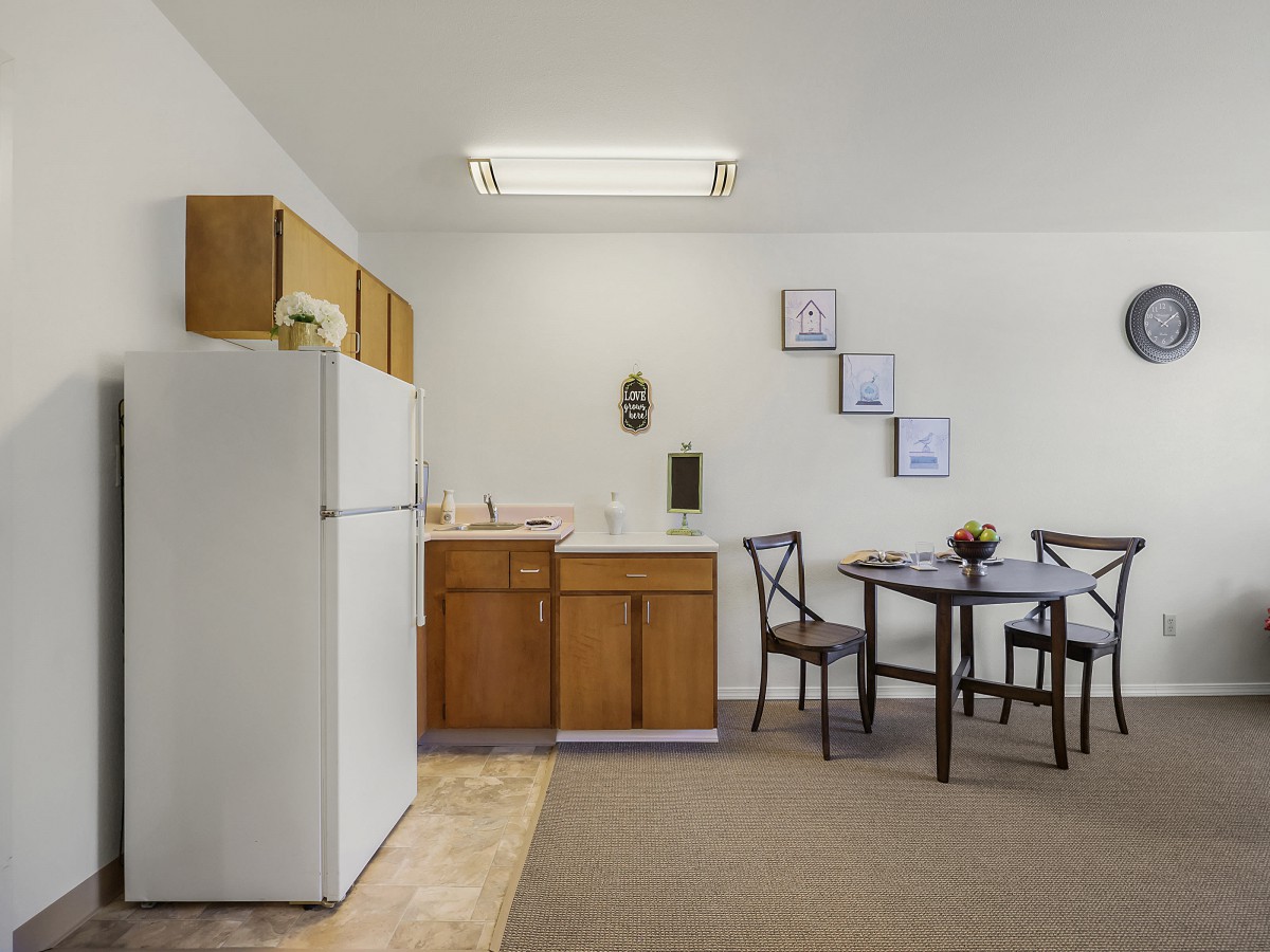 A simple kitchen and dining area with a refrigerator, wooden cabinets, and a small dining table with chairs.