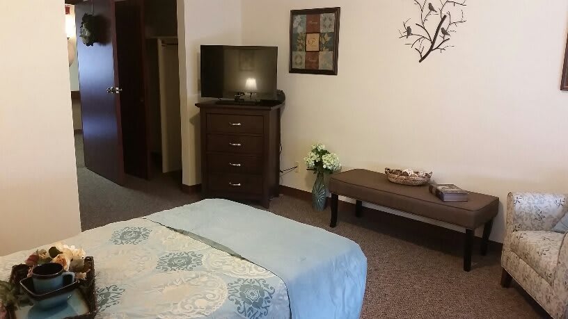 A cozy bedroom featuring a TV on a dresser, a neatly made bed, and a decorative bench with flowers.