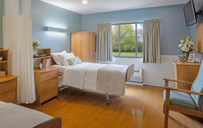 A hospital room with a single bed, wooden furniture, and a large window with curtains.