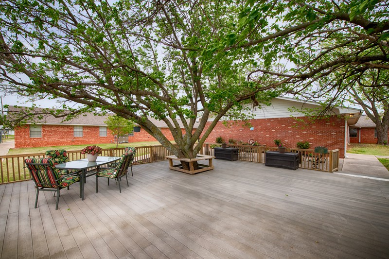 A spacious wooden deck with a large tree in the center, surrounded by outdoor seating and a brick wall backdrop.