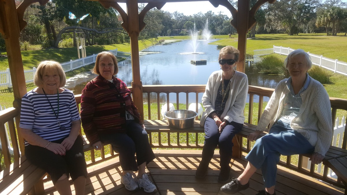 A group of people sit in a gazebo overlooking a lake with a fountain on a sunny day.
