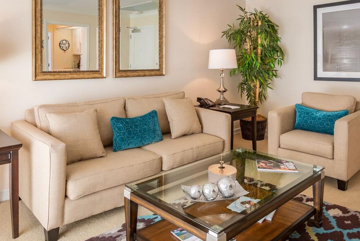 A cozy living room with beige sofas and teal cushions, featuring a glass coffee table and elegant decor.