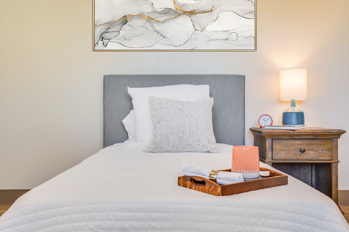 A neatly arranged bedroom with a gray headboard, a pillow, and a tray on the bed beside a lamp on the nightstand.