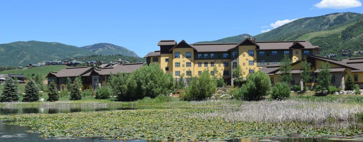 A scenic view of a building surrounded by lush greenery and a pond, with mountains in the background under a clear blue sky.