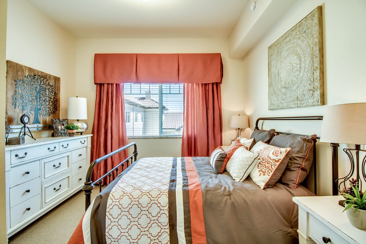 A cozy bedroom with stylish decor and a large window, featuring warm-toned curtains and bedding.