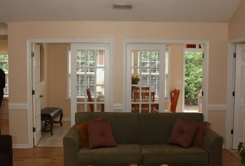 A cozy living room with a green couch facing toward the dining area, featuring large windows and warm lighting.