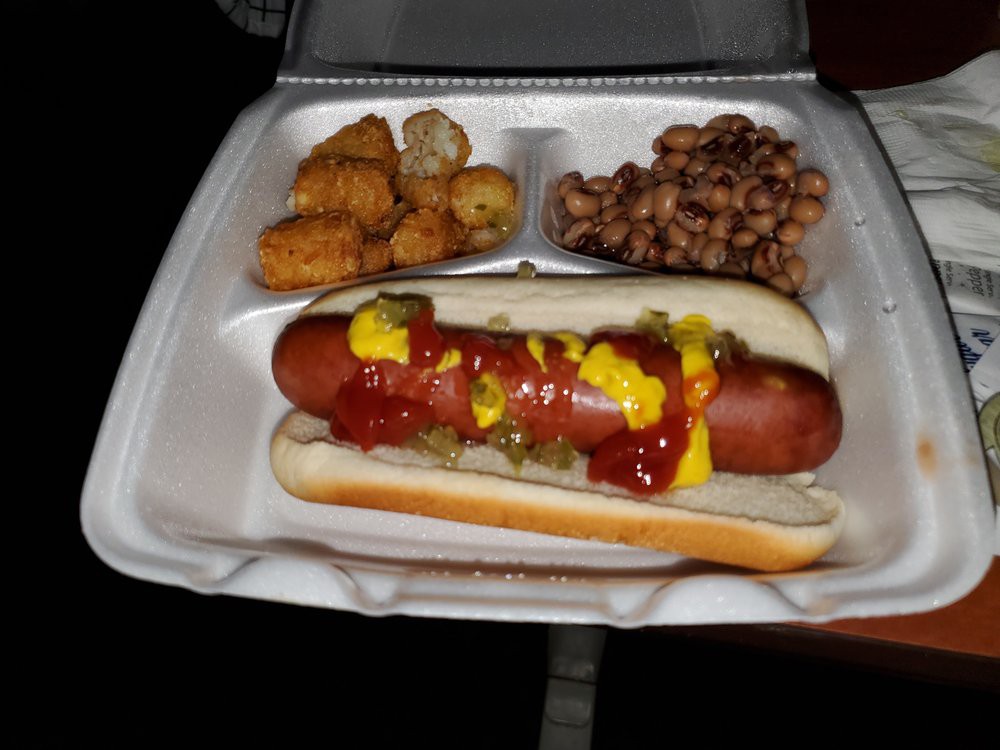 A meal consisting of a hot dog with ketchup and mustard, accompanied by tater tots and baked beans in a takeout container.