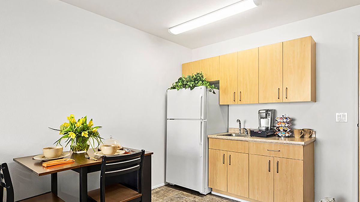 A compact kitchen area with light wooden cabinets, a white refrigerator topped with plants, and a small dining table decorated with flowers.