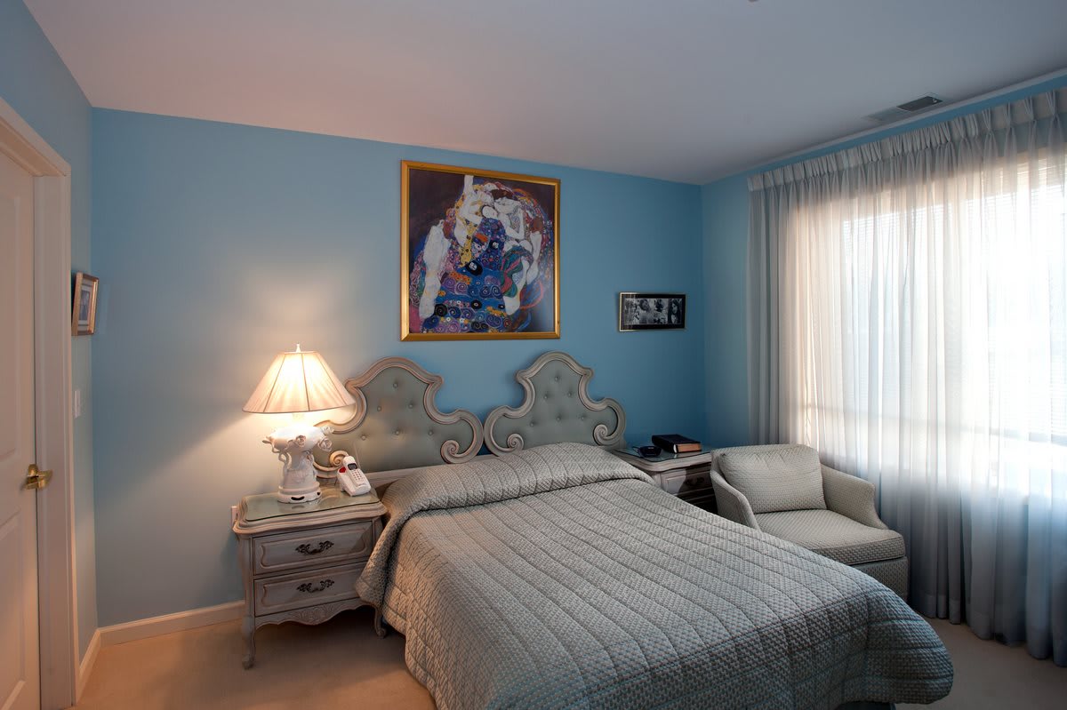 A cozy bedroom with blue walls, featuring a bed, nightstands, and a chair, adorned with a decorative painting.