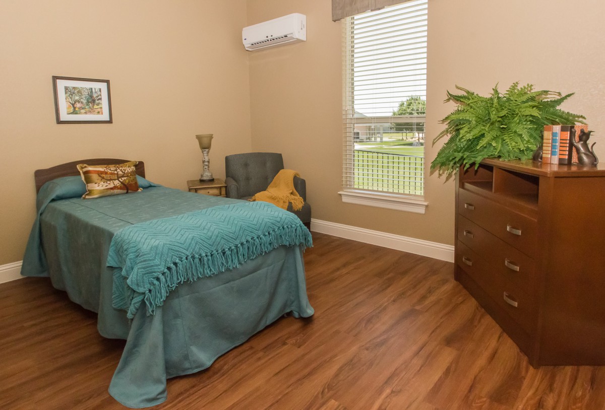 A cozy, minimalist bedroom featuring a single bed with teal bedding, wooden flooring, and a large plant near the window.